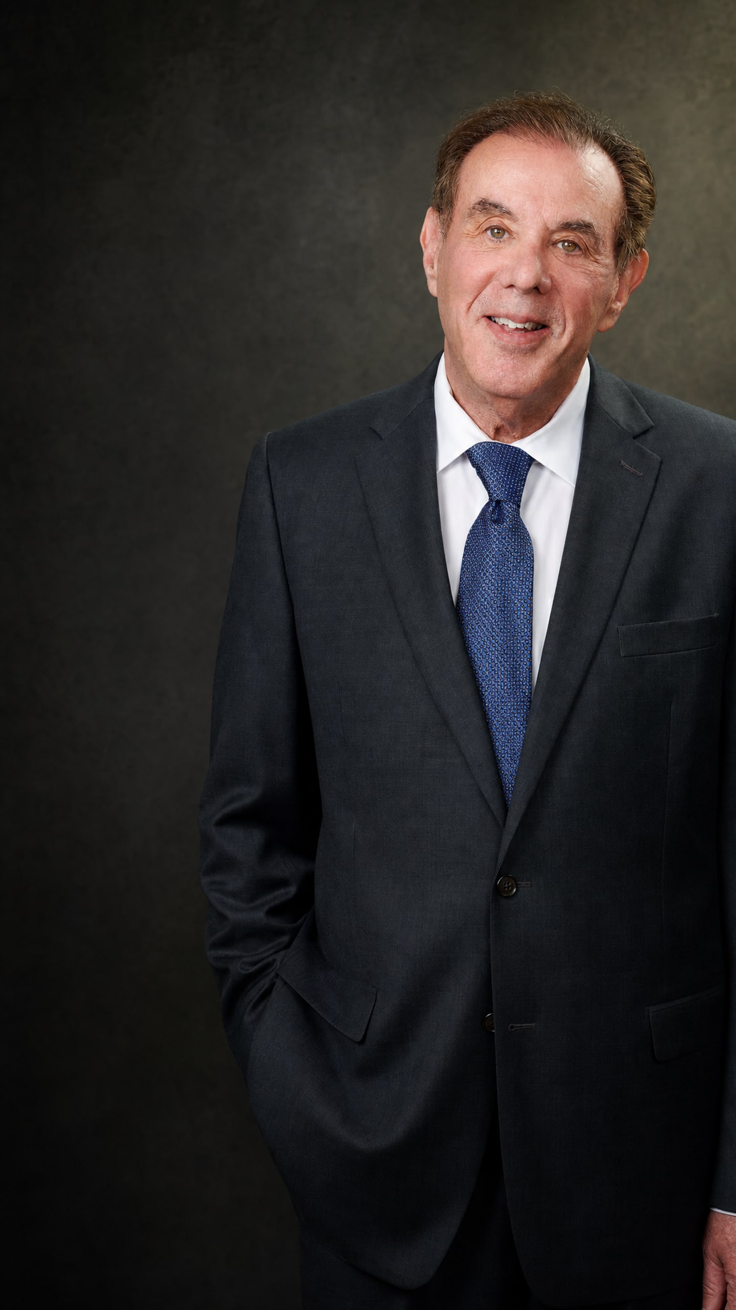 William Turkish in a charcoal suit against a dark olive background.